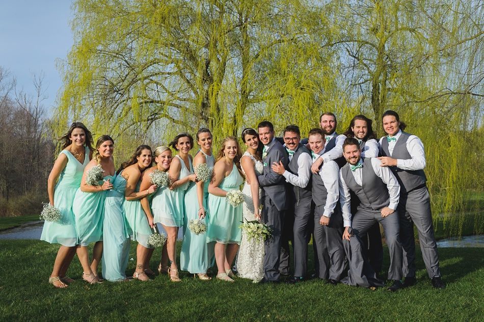 Wedding party posing outdoors. Bridesmaids in teal dresses, groomsmen in gray vests, couple center. Willow tree backdrop.
