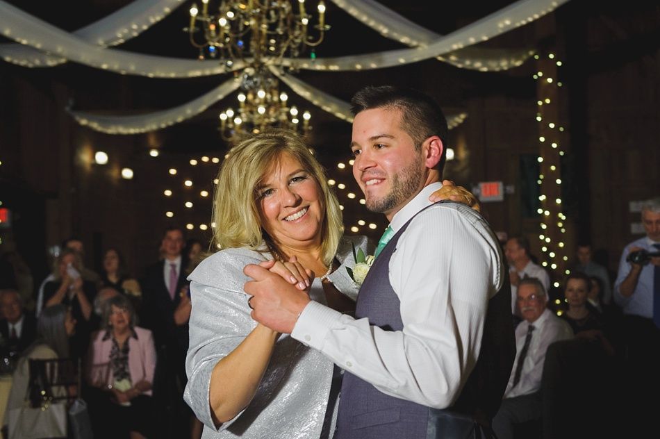 A man and woman dance at a wedding. The man smiles and wears a vest. The woman wears a silver jacket.