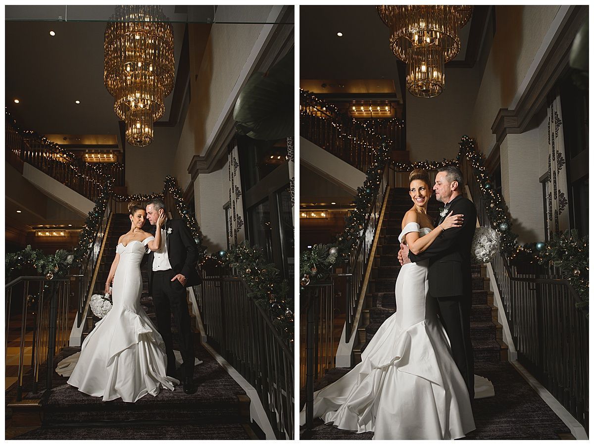 Bride and groom embrace on staircase, beneath chandelier, decorated for an event.