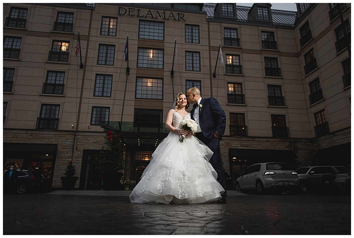 Bride and groom in front of a hotel, she in white dress, he in suit. Hotel exterior lit, overcast.