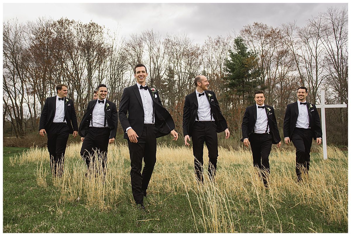 Six men in tuxedos walking through a field, smiling, with a backdrop of trees and a cloudy sky.