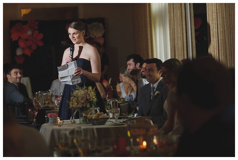 Bride with arms up, making a funny face, groom looking at his hand, cake cutting in background.