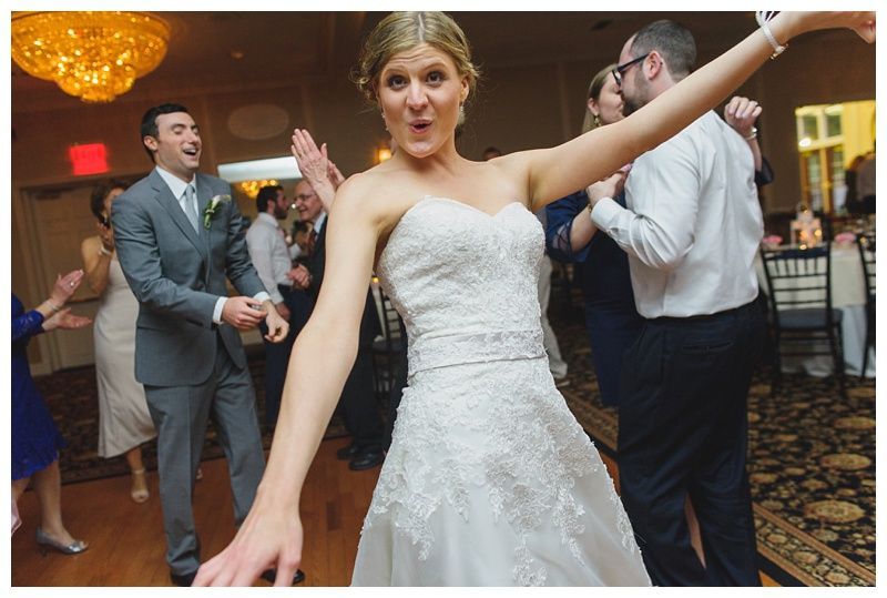 Bride with arms up, making a funny face, groom looking at his hand, cake cutting in background.