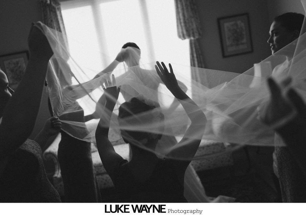 Bridesmaids helping bride with veil in a sunlit room. Blurred figures with outstretched arms. Black and white.