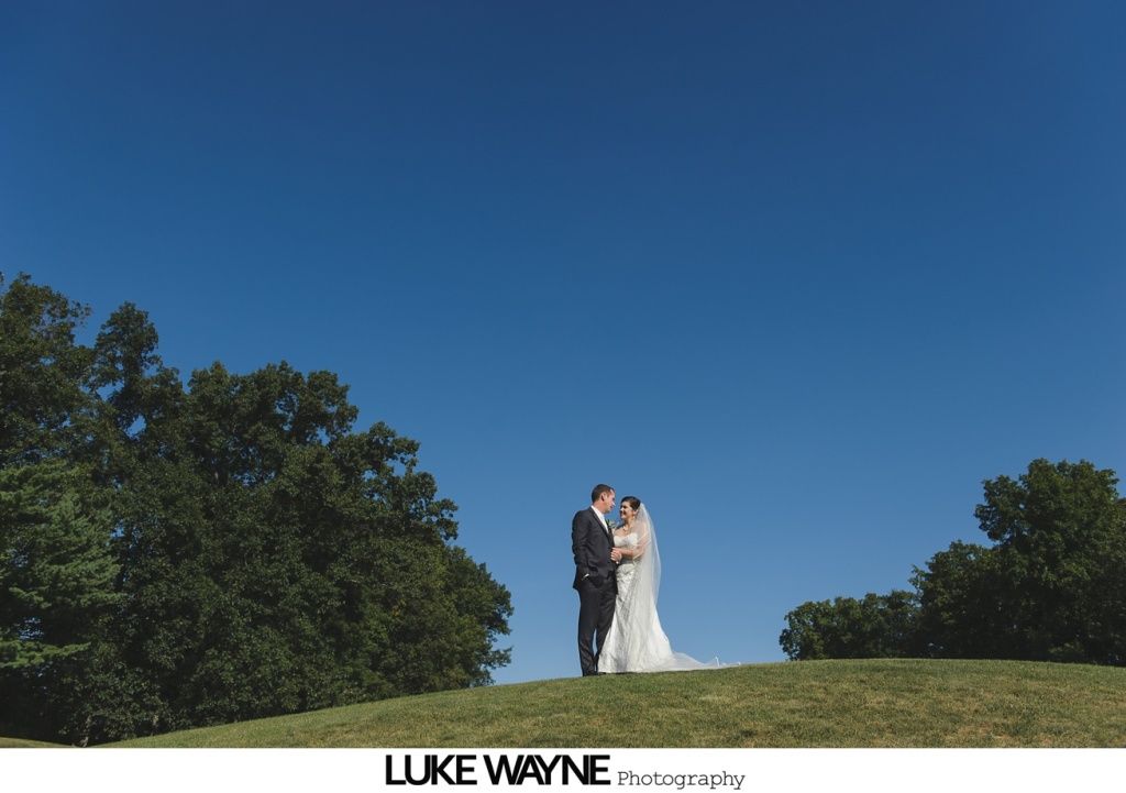 Bride and groom standing on a grassy hill, embracing under a bright blue sky.