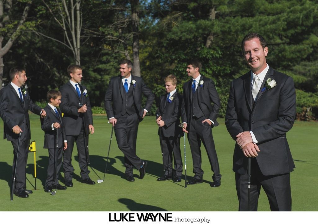 Groomsmen in dark suits on a golf course, holding golf clubs. The groom smiles, others look on.