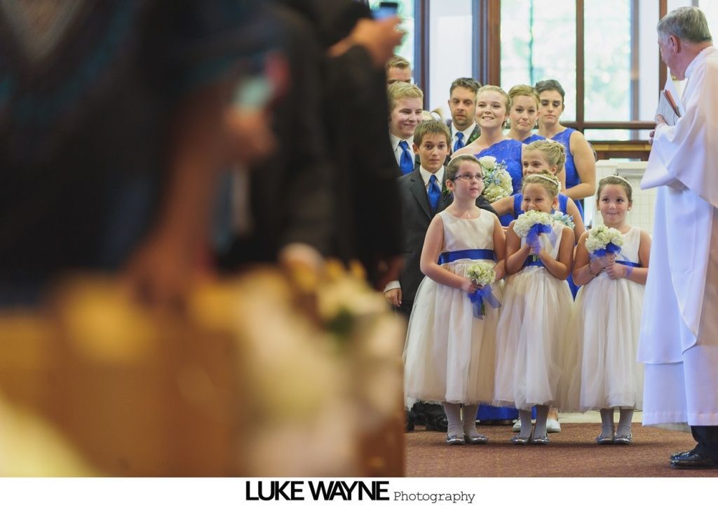 Wedding ceremony: Flower girls in white dresses, bridesmaids in blue, groom and wedding party in suits, church setting.