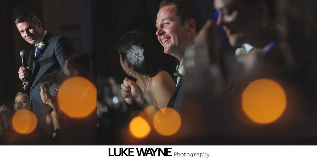 Man giving a speech at a wedding, smiling couple and guests in the blurred background, lit by warm bokeh.