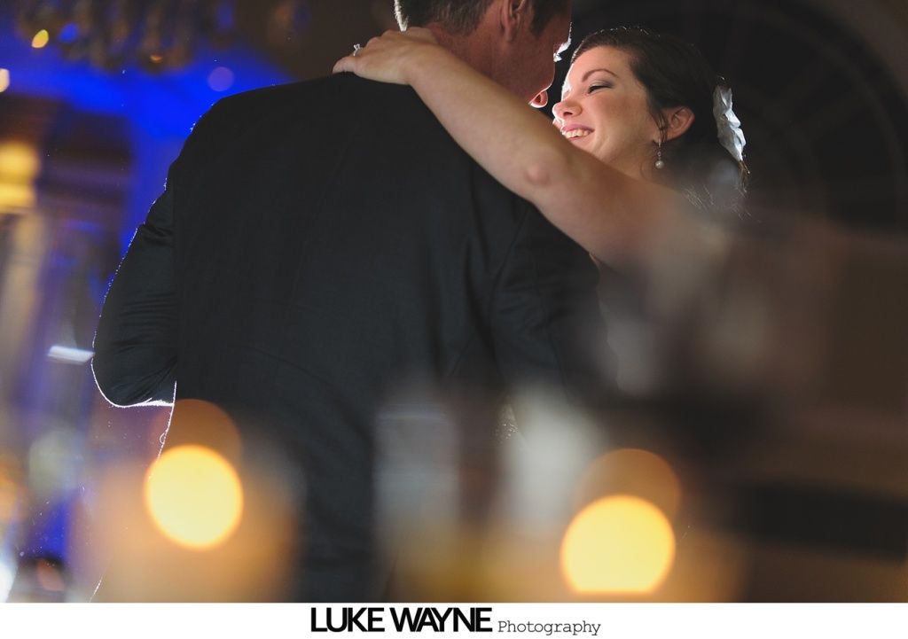 Couple dancing at a wedding, woman smiles, blue and yellow lights in background.