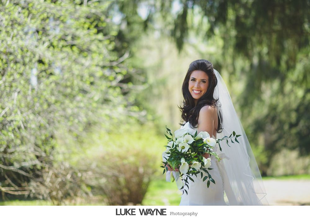 Bride smiles holding a bouquet, standing outside with trees in the background.