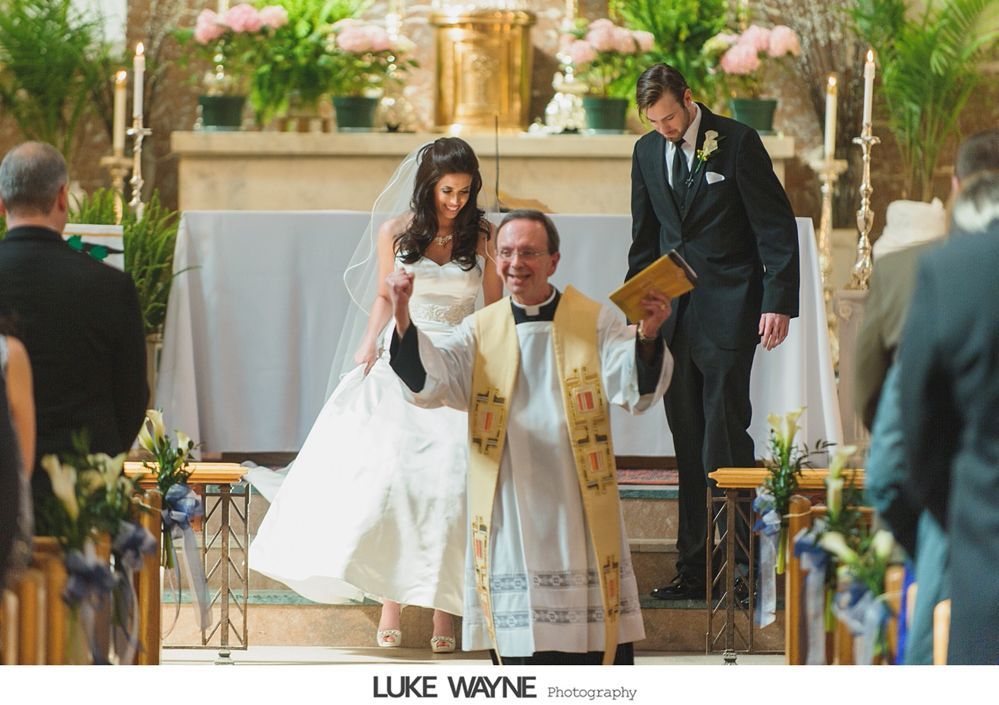 Bride and groom with priest at altar, smiling after wedding ceremony. Church interior with floral decorations.