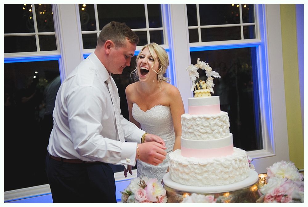 Bride with arms up, making a funny face, groom looking at his hand, cake cutting in background.
