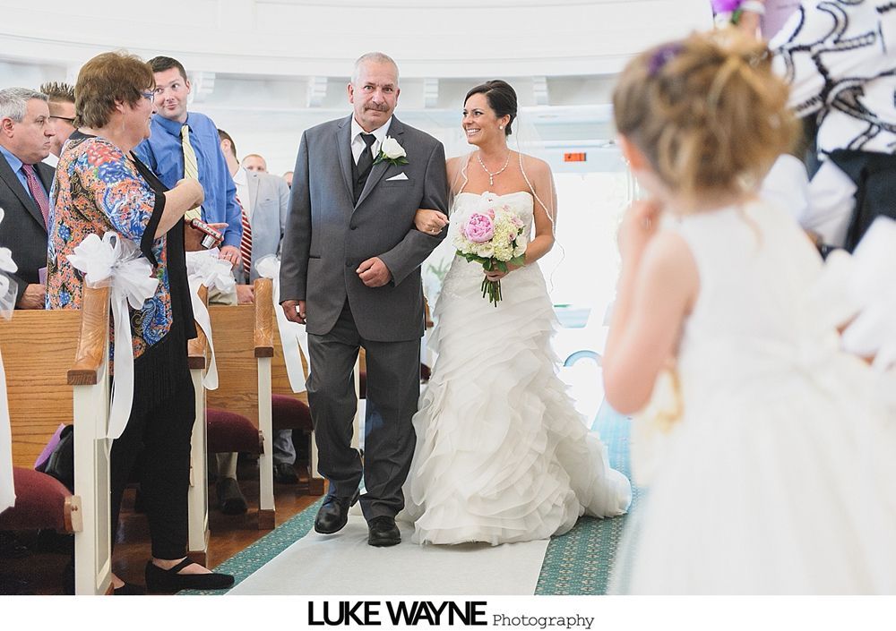 Newlyweds walking down the aisle after a wedding ceremony, guests clapping. Glass enclosed building, white paper lanterns overhead.