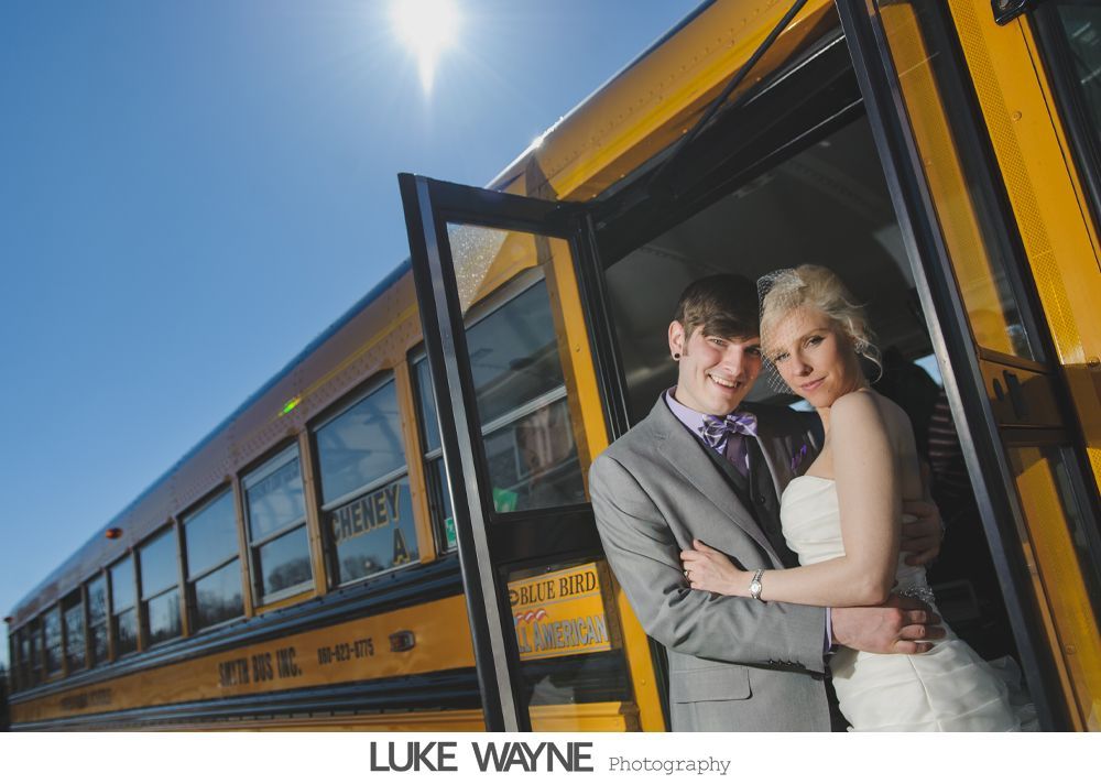 Couple in wedding attire embrace by open school bus door under a sunny sky.