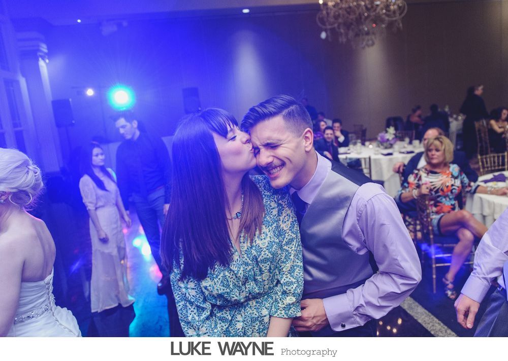Woman kisses smiling man's cheek at a party; blue lights and wedding guests in background.