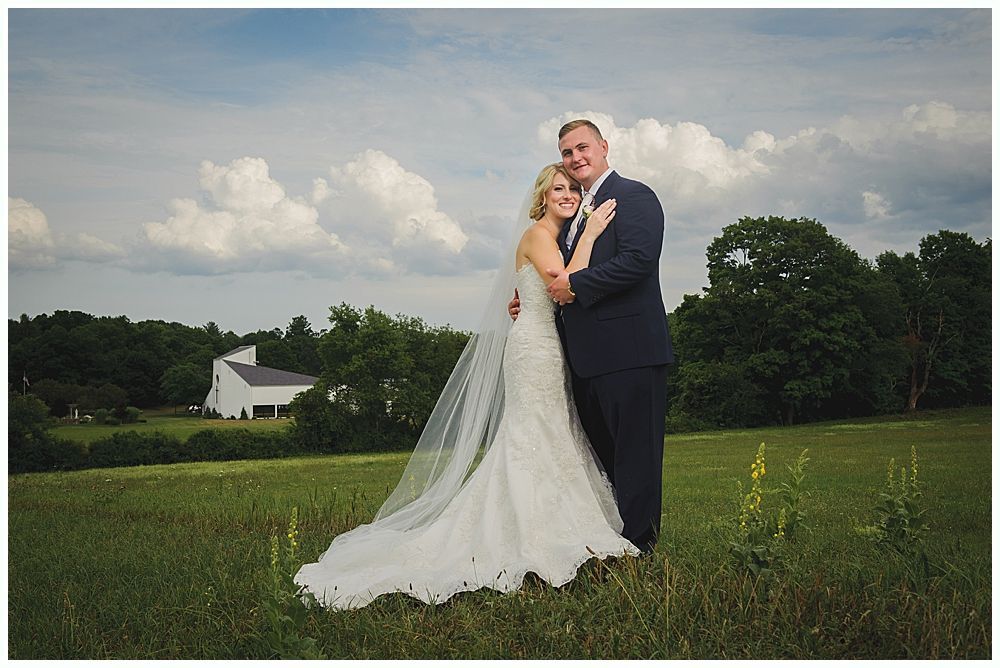 Bride and groom embrace in a field, a white building in the distance, cloudy sky overhead.
