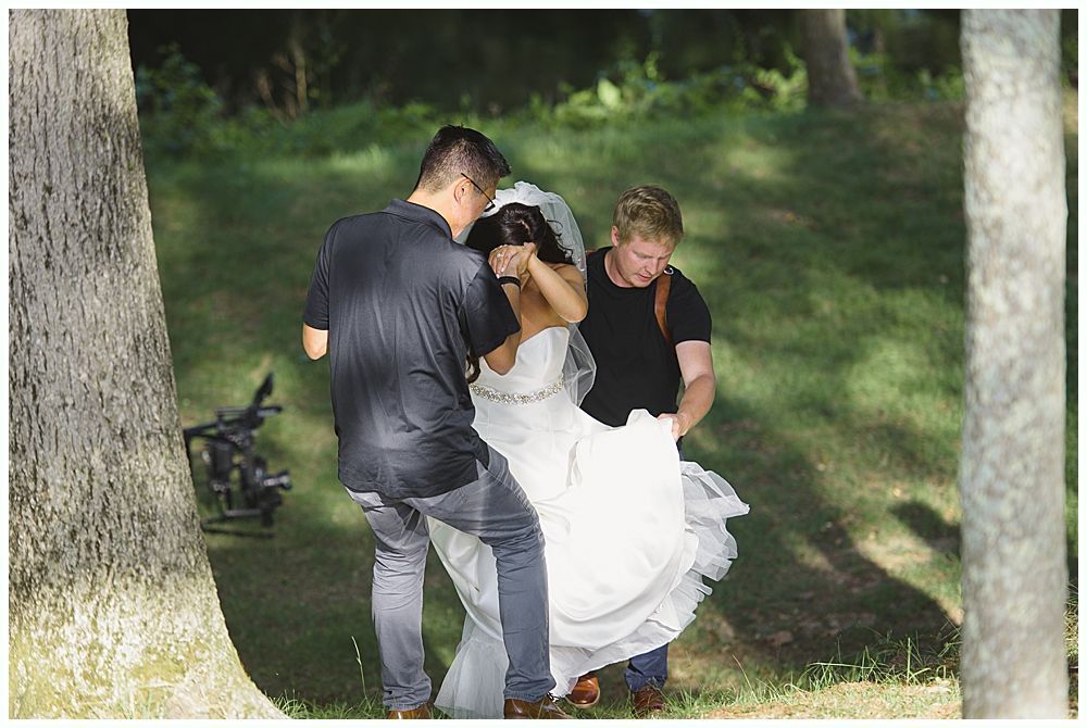 Two men help a bride with her dress as she walks on grass near trees.