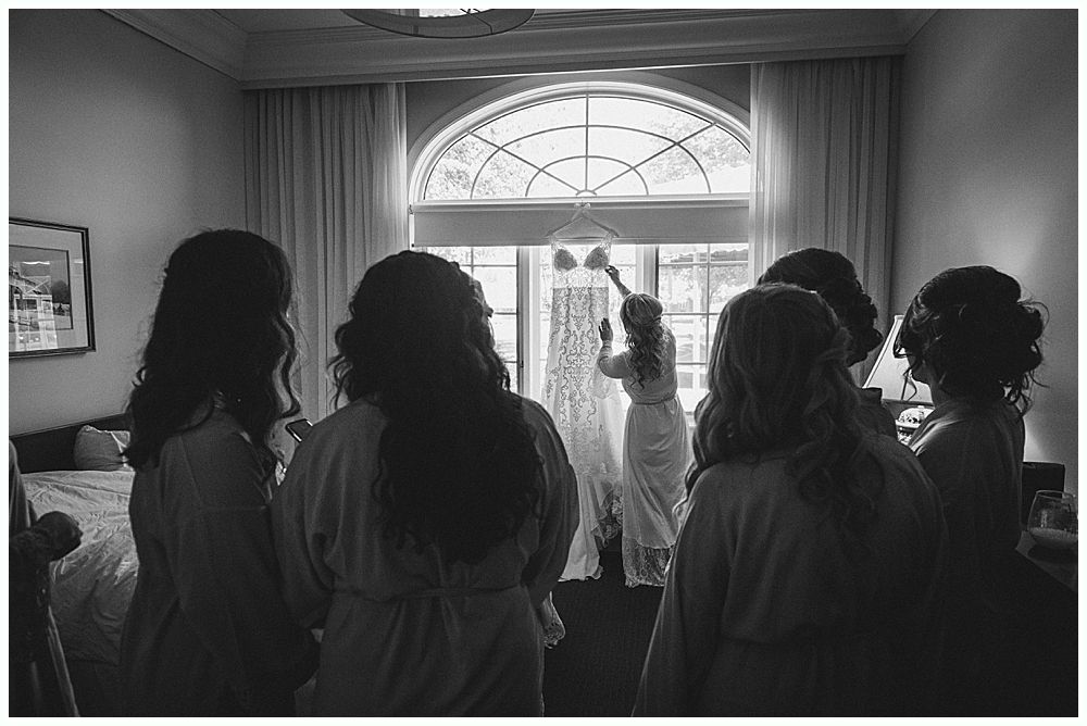 Bridesmaids watch bride adjust wedding dress hanging in front of a window. Black and white.