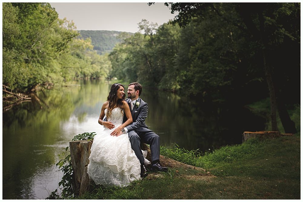 Bride and groom embrace by a river, smiling. Bride in white dress, groom in suit, surrounded by trees.
