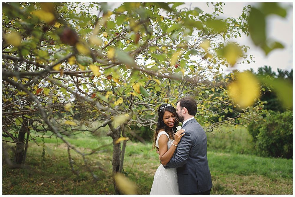 Couple embraces outdoors near a tree with fall leaves; woman smiles, man kisses her head.