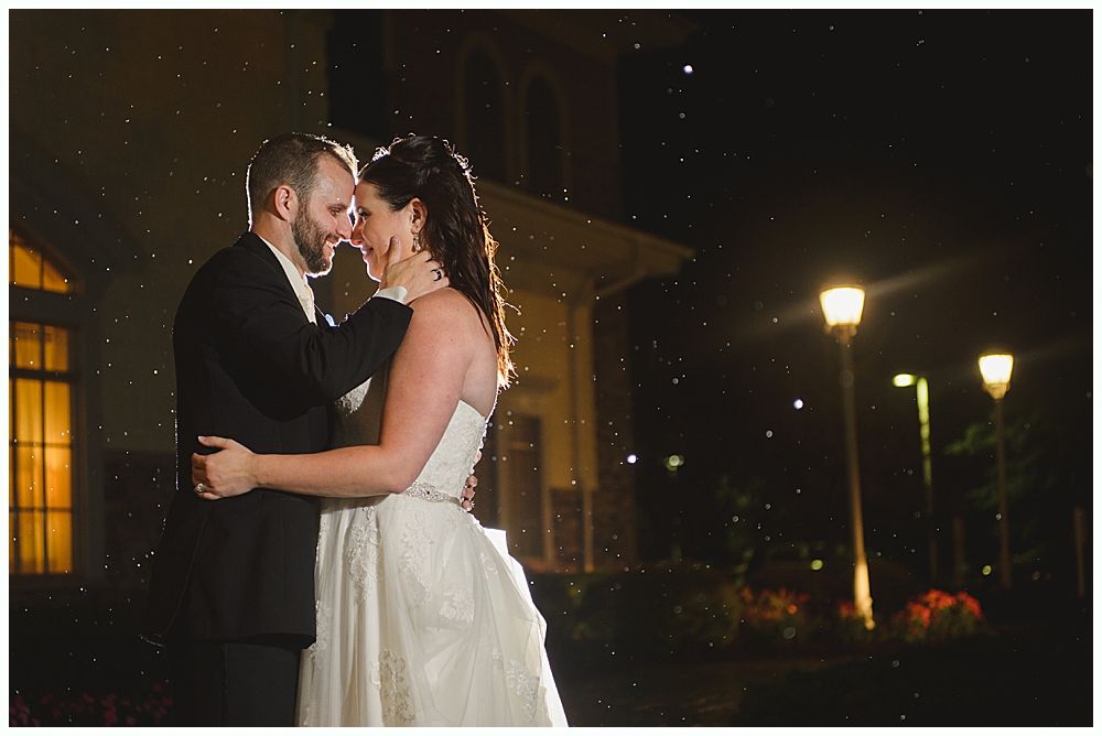Bride and groom embracing, lit by lights at night, outside a building with arched windows.