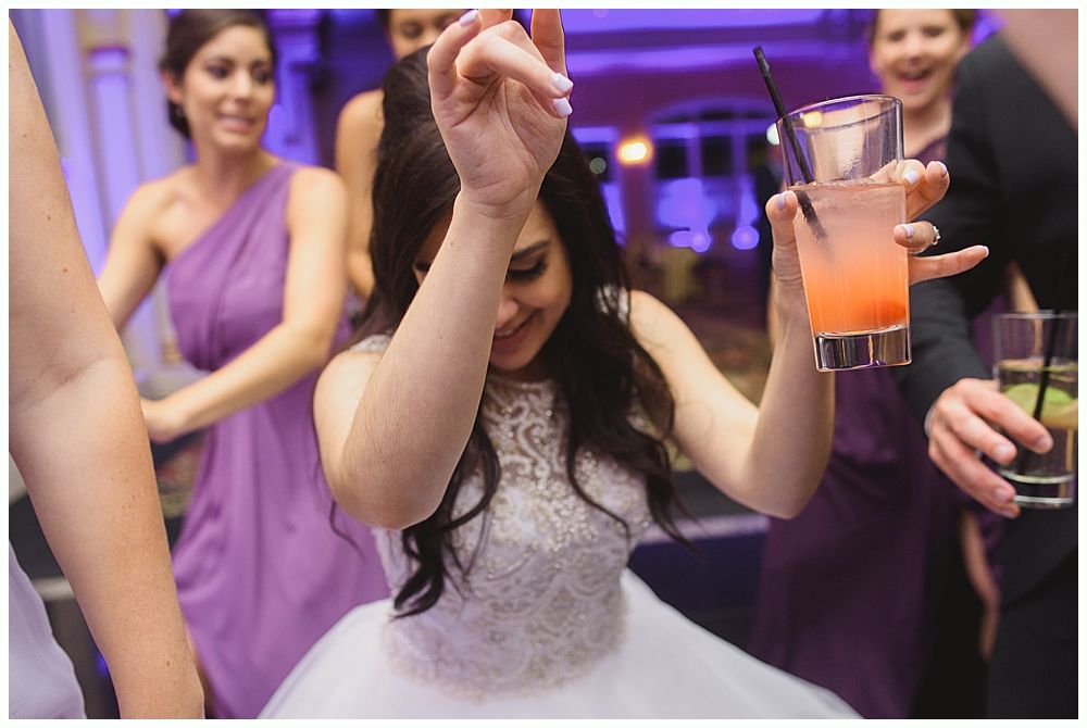 Bride dancing at a party, holding a drink, wearing a white dress. Bridesmaids in purple dresses smile in the background.