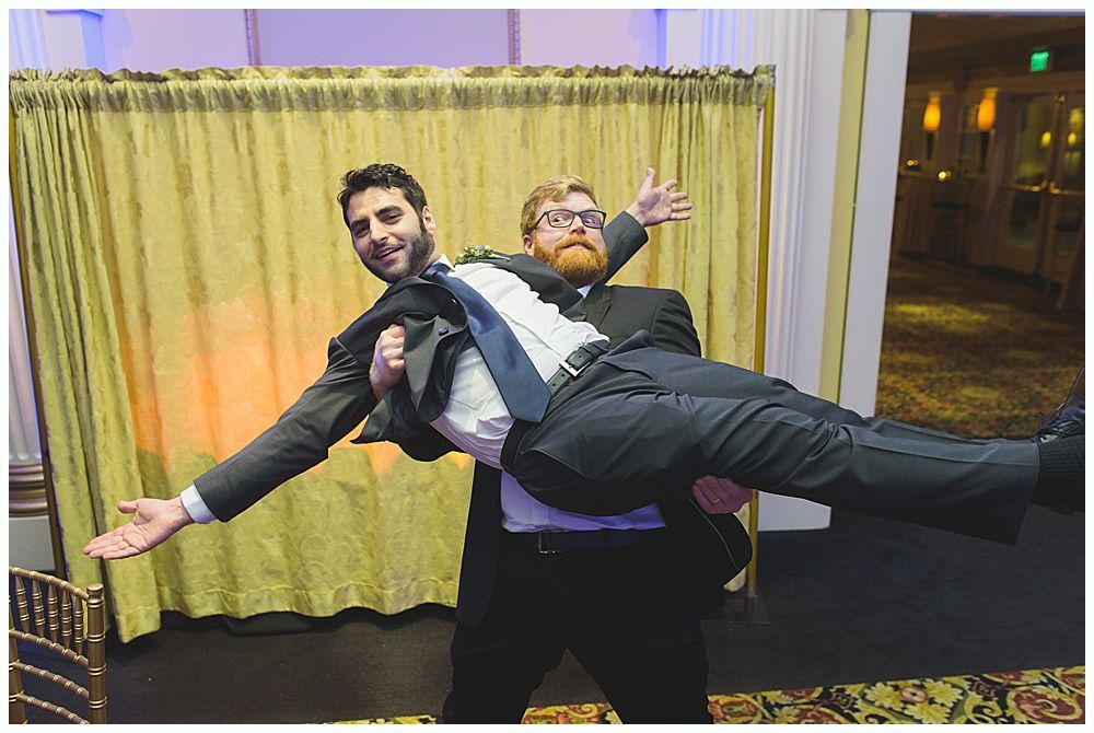 Two men in suits, one carrying the other. Background with gold curtains and a carpeted room.