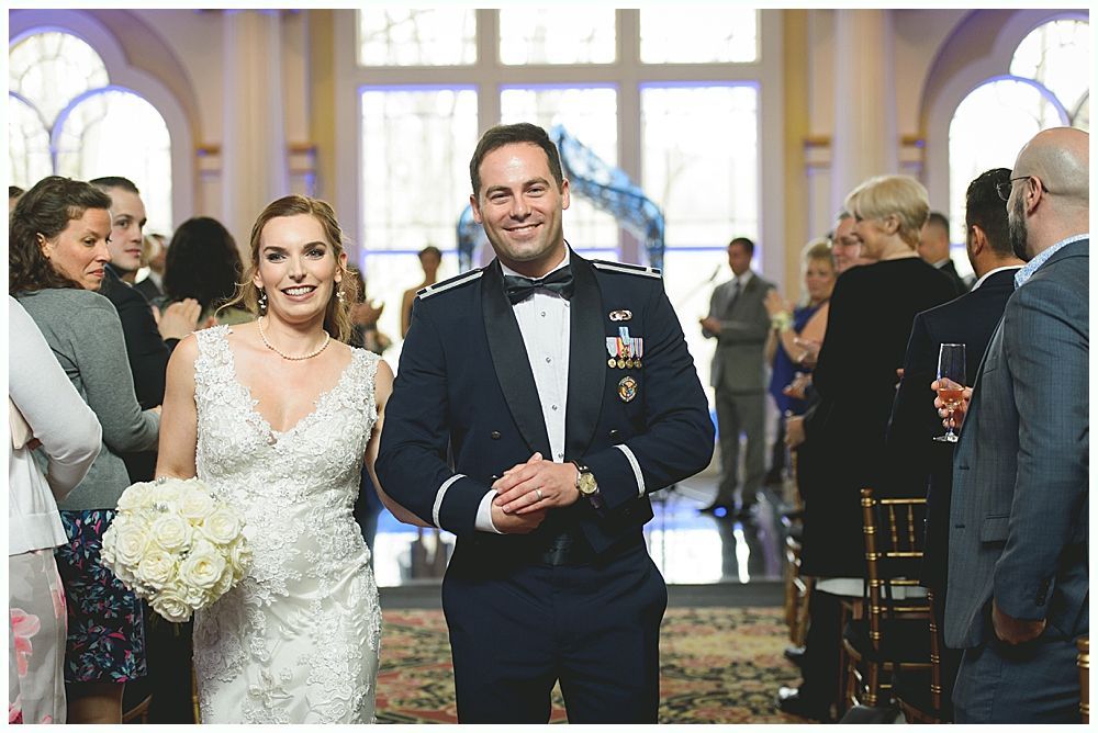 Bride and groom smiling, walking down aisle after wedding. Groom in military uniform, bride in white gown.