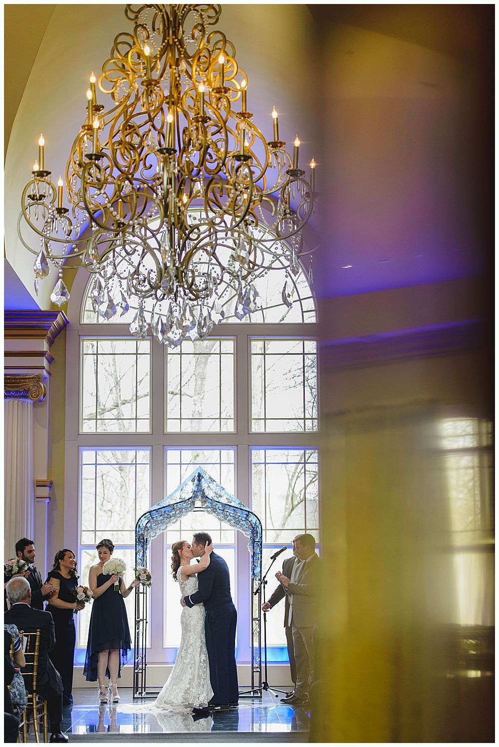 Couple kissing under floral arch during wedding ceremony, ornate chandelier overhead.