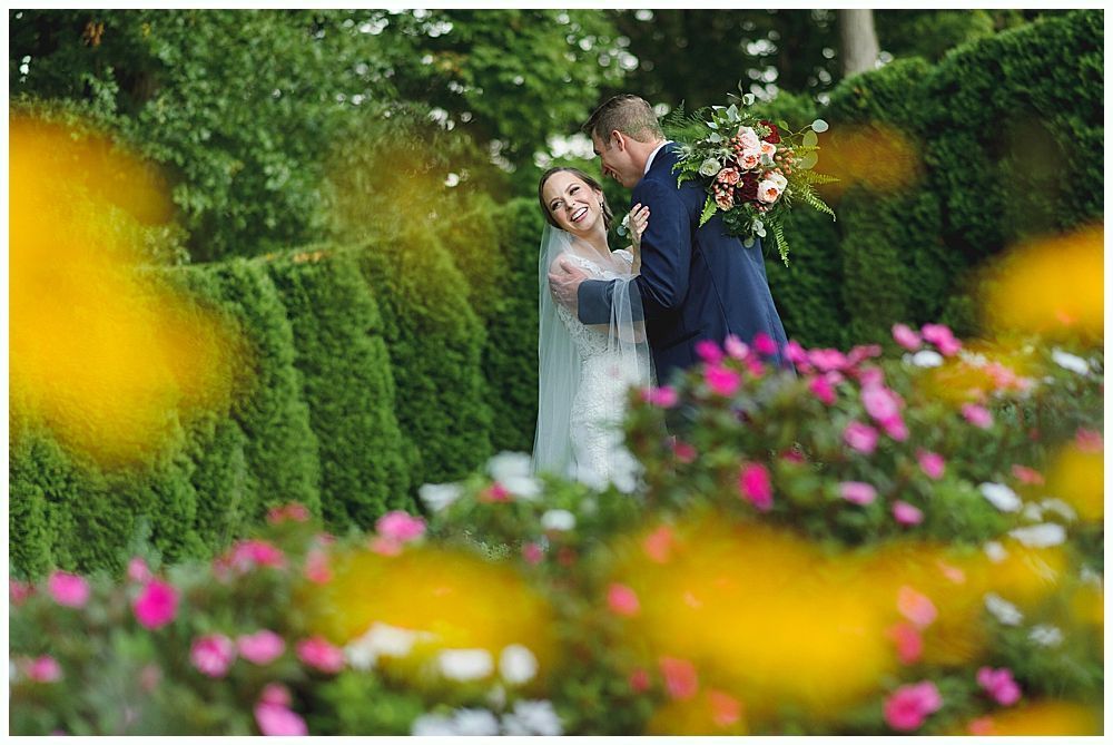 Bride and groom embrace in a garden. The bride smiles, holding a bouquet. Green hedges and flowers surround them.