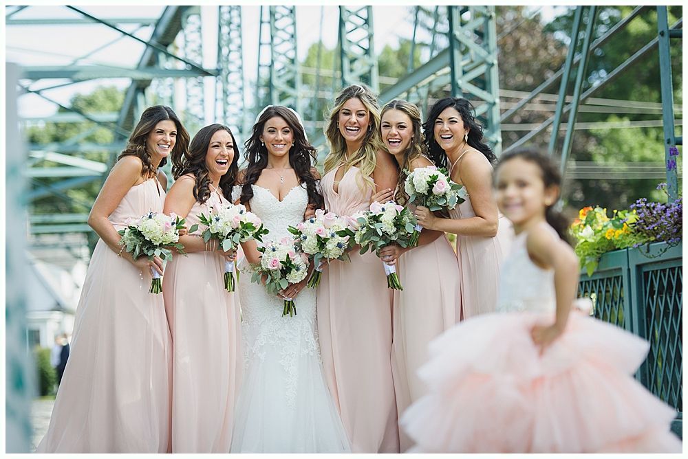 Bride and bridesmaids in blush gowns holding bouquets on a bridge; flower girl in foreground.
