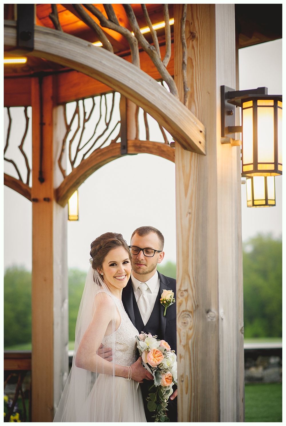 Bride and groom smiling, posing under a wooden archway with lights. Bride holds a bouquet.