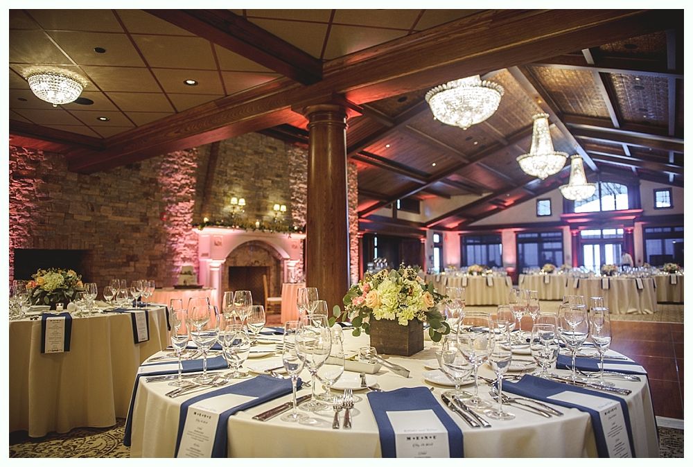 Formal dining room, round tables set with blue accents, lit by chandeliers. Wooden beams and stone fireplace in background.