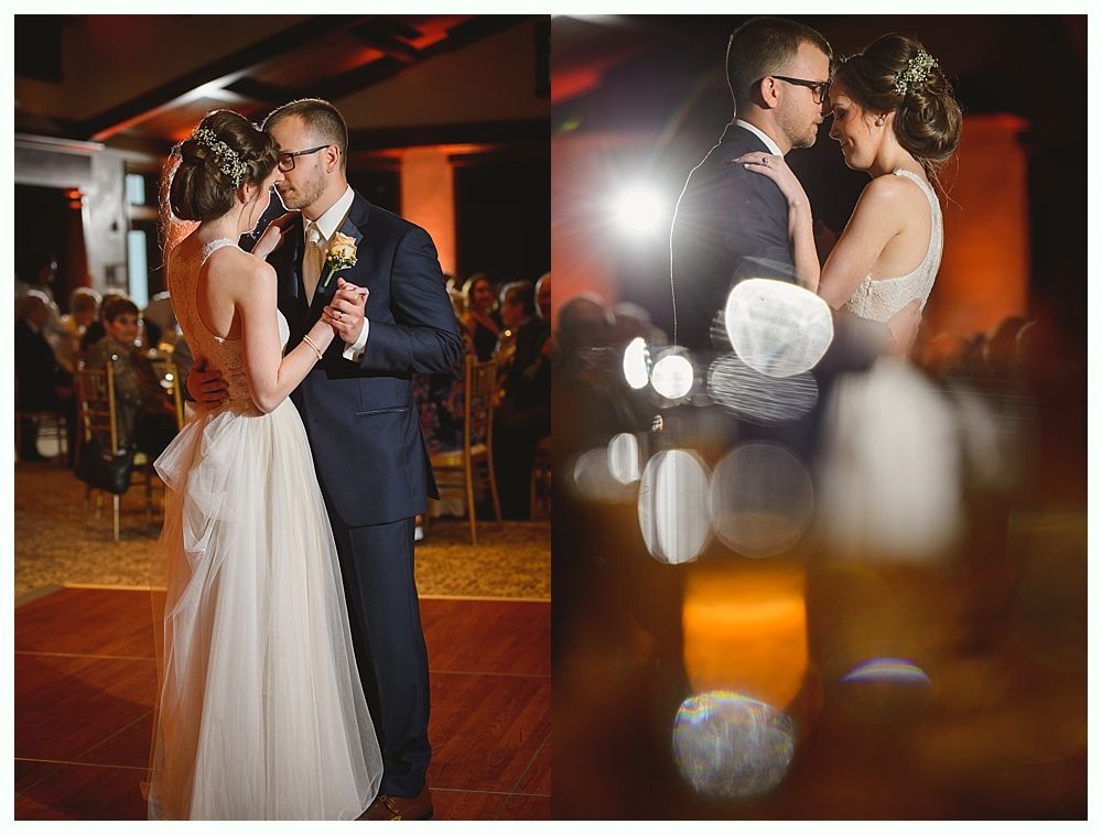 Wedding couple dancing in a ballroom, she in a white dress, he in a blue suit. Warm lighting and guests in the background.