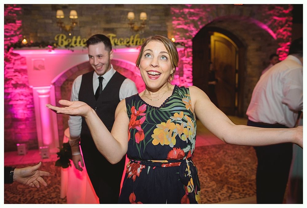 Woman dancing excitedly at a wedding reception with open arms; man in background smiles. Pink lighting.