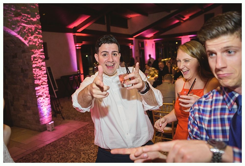 Man points a finger with cocktail in hand, smiling at the camera, flanked by laughing friends in a warmly lit party setting.