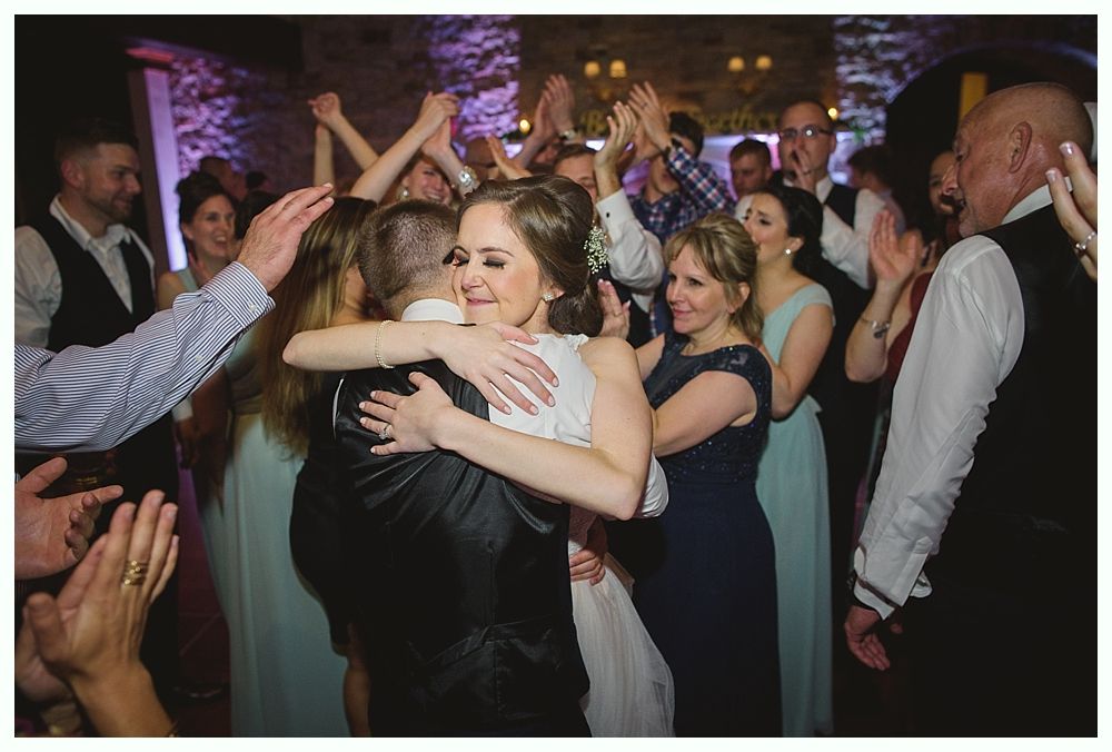 Bride and groom embrace, surrounded by cheering guests at a wedding reception.