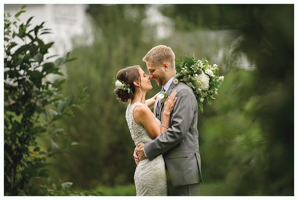 Wedding couple embracing outdoors, woman in white dress, man in gray suit, greenery background.