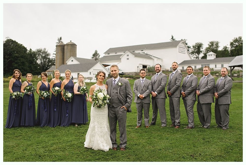 Wedding party posing on a grassy field in front of a white barn. Bride and groom in the center, bridesmaids in navy, groomsmen in gray suits.