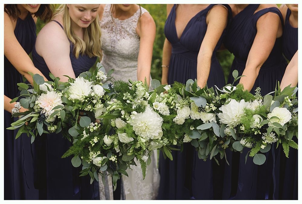Bridesmaids in navy dresses holding a long floral arrangement of white flowers and greenery outdoors.