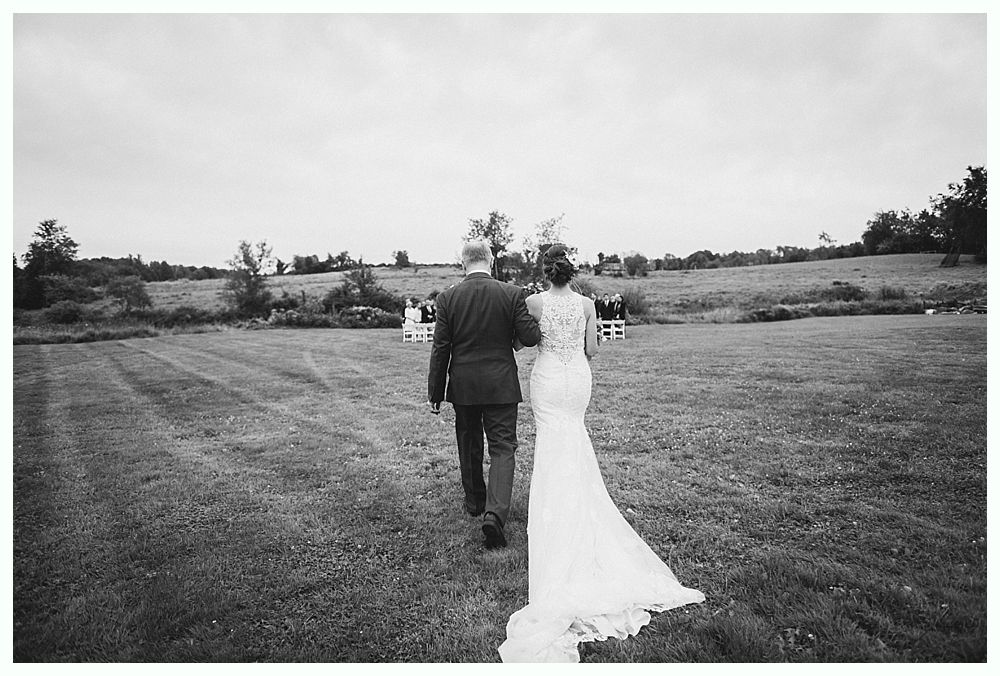 Bride and her father walk down a grassy aisle towards a ceremony, back view, black and white photo.