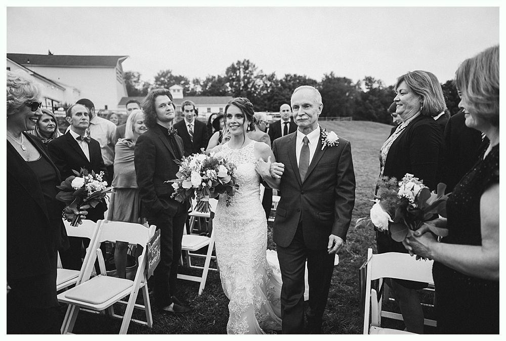 Bride and father walk down aisle, guests watching. Wedding ceremony outdoors. Black and white.