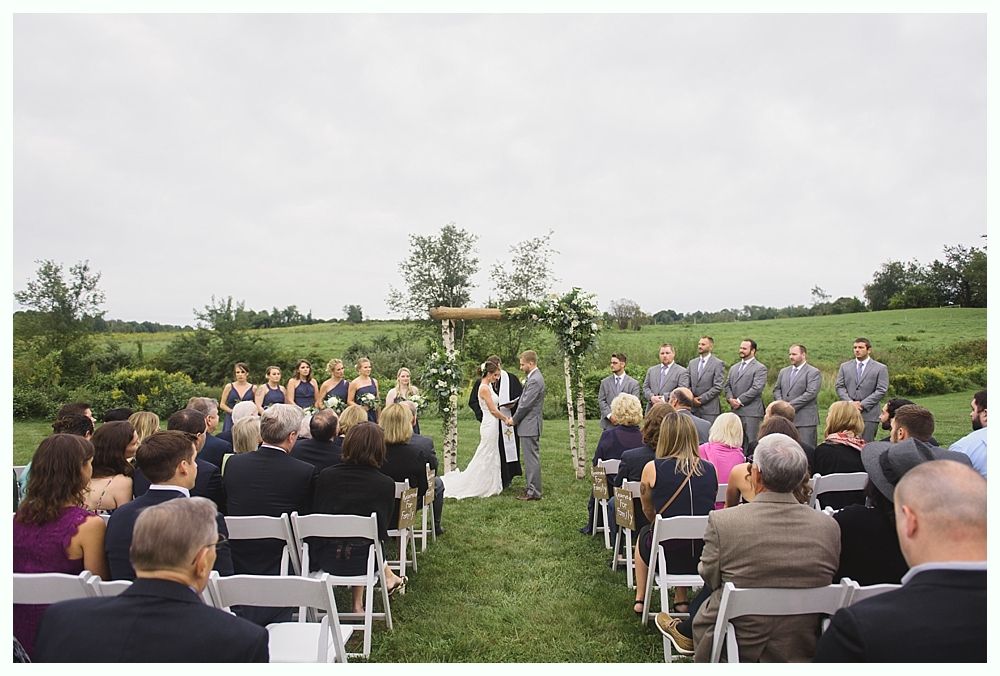 Wedding ceremony outdoors; couple under arch, wedding party and guests seated, grassy field.