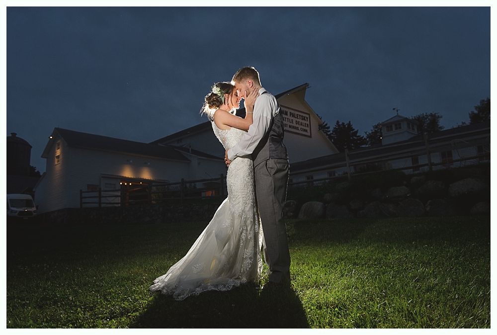Couple embracing in front of a white barn at dusk. They are illuminated by light.