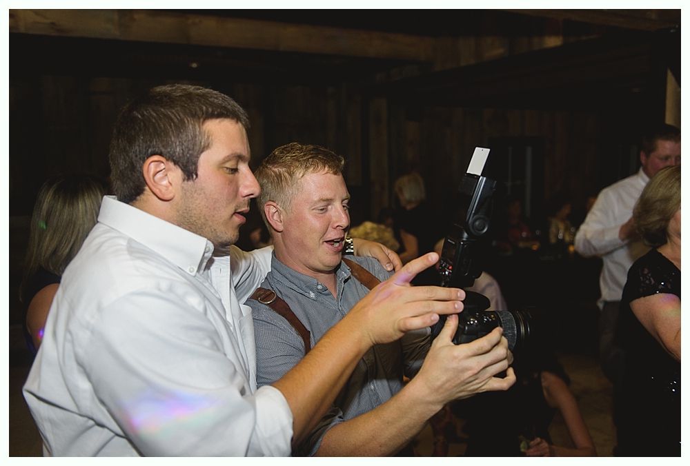 Two men examining a black object, possibly a camera, in a dimly lit room with other people.