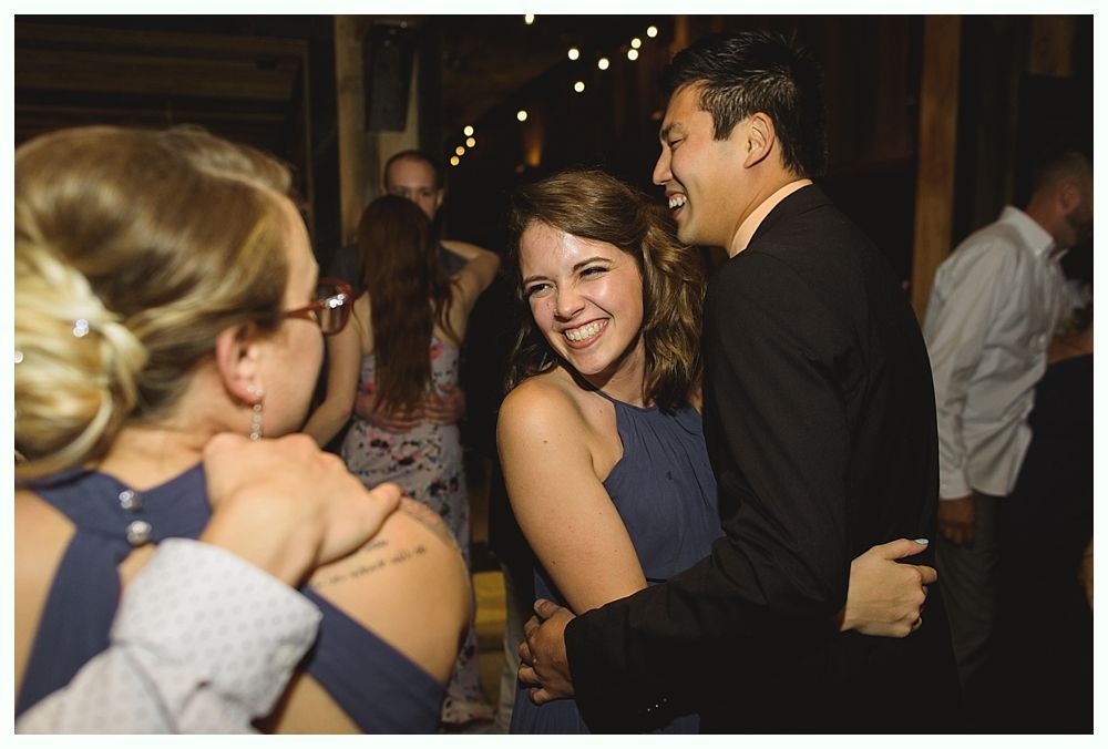 People dancing at a party; woman in blue dress laughs, hugs man in black suit. Dim lighting, string lights.