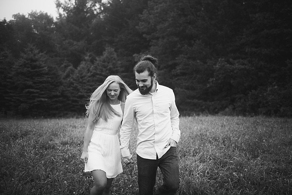 Couple holding hands, walking through a field. Man with beard, ponytail; woman in white dress.