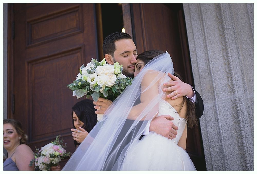 Newlyweds embrace outside a building. Bride in white dress and veil, holding bouquet; groom in suit.