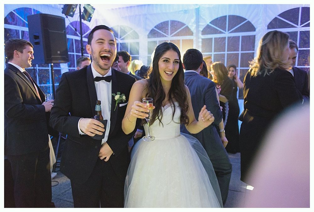 Bride and groom dancing at a wedding reception, laughing and holding drinks under a tent.