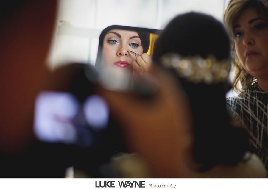 Woman having makeup applied, looking in mirror. Another person holds a phone and woman looks on.